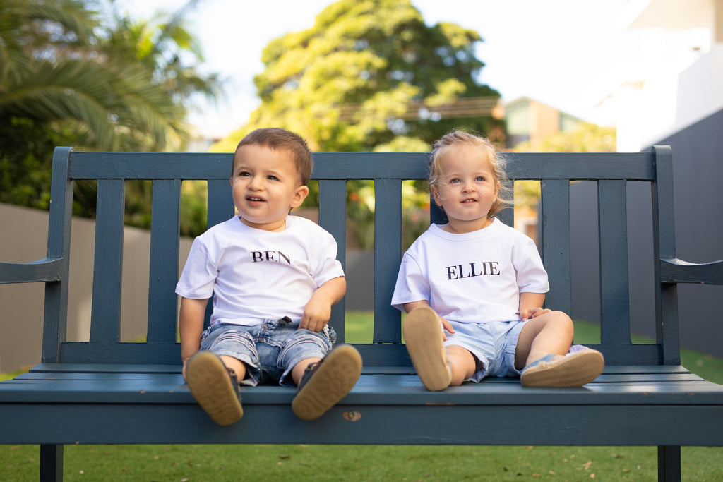Two toddlers sitting on a seat. Both wearing a white shirt, one with the name Ben and the other with the name Ellie. 